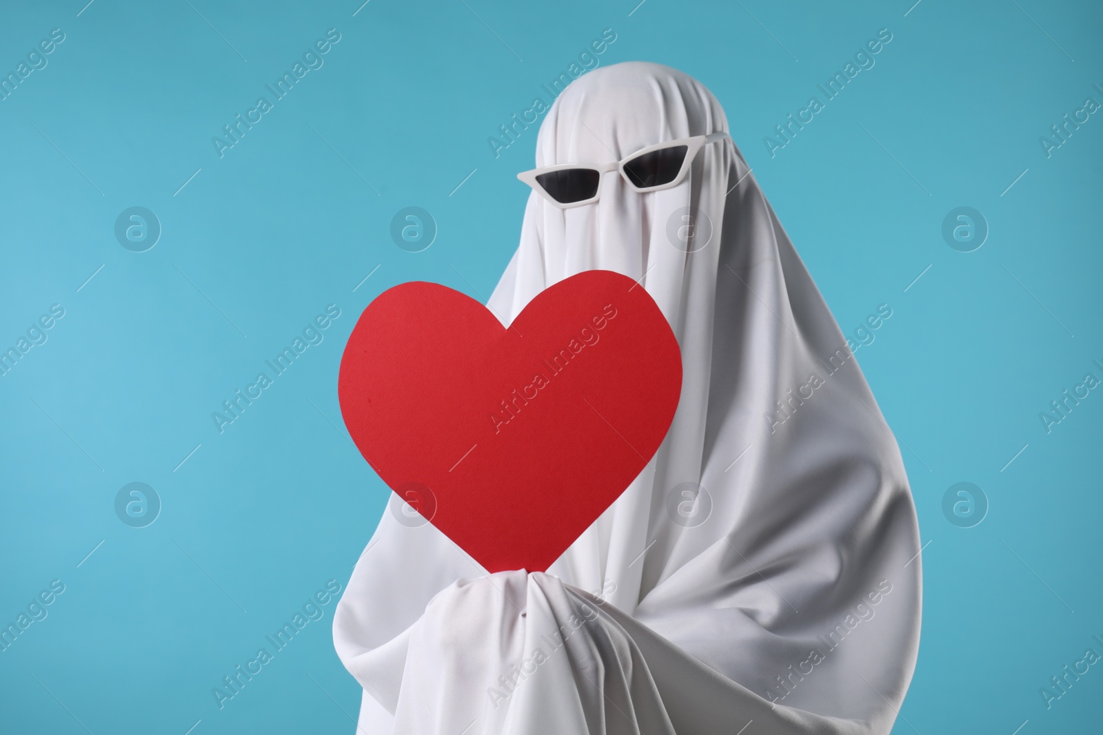 Photo of Cute ghost. Person covered with white sheet in sunglasses holding red heart on light blue background