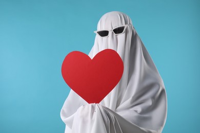 Photo of Cute ghost. Person covered with white sheet in sunglasses holding red heart on light blue background