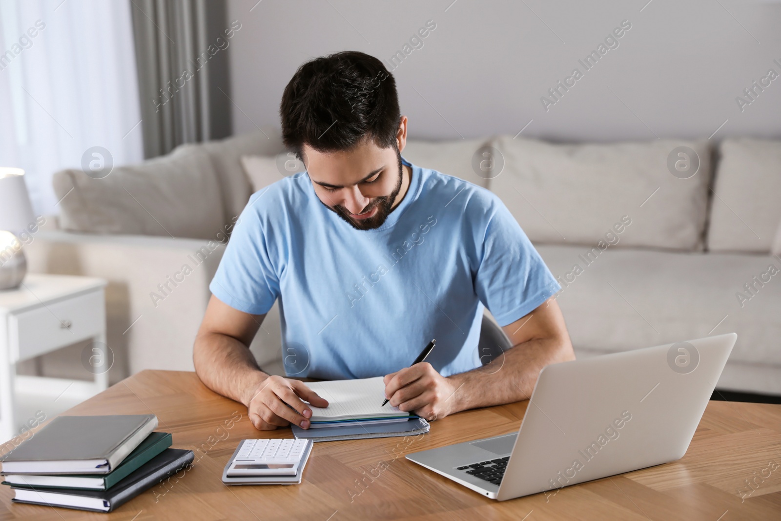 Photo of Young man writing down notes during webinar at table in room