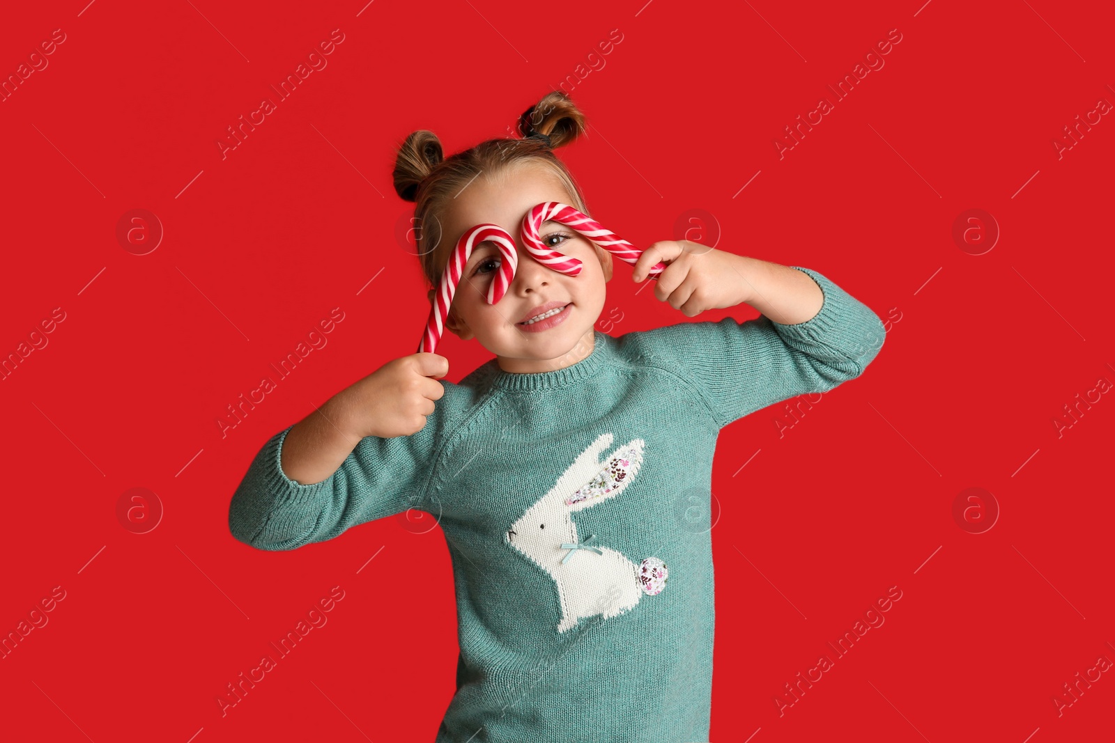 Photo of Cute little girl in knitted sweater holding candy canes on red background. Celebrating Christmas