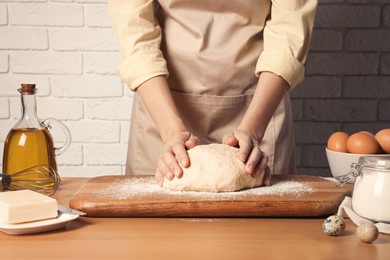 Photo of Woman kneading dough at wooden table near white brick wall, closeup