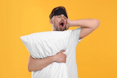 Photo of Sleepy man with pillow and sleep mask yawning on yellow background. Insomnia problem