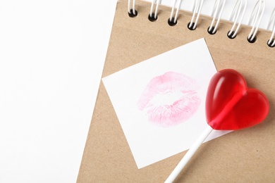 Photo of Heart shaped lollipop, notebook and paper note with lipstick kiss on white background, flat lay. Valentine's day celebration