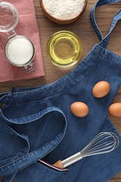 Denim apron with kitchen tool and different ingredients on wooden table, flat lay