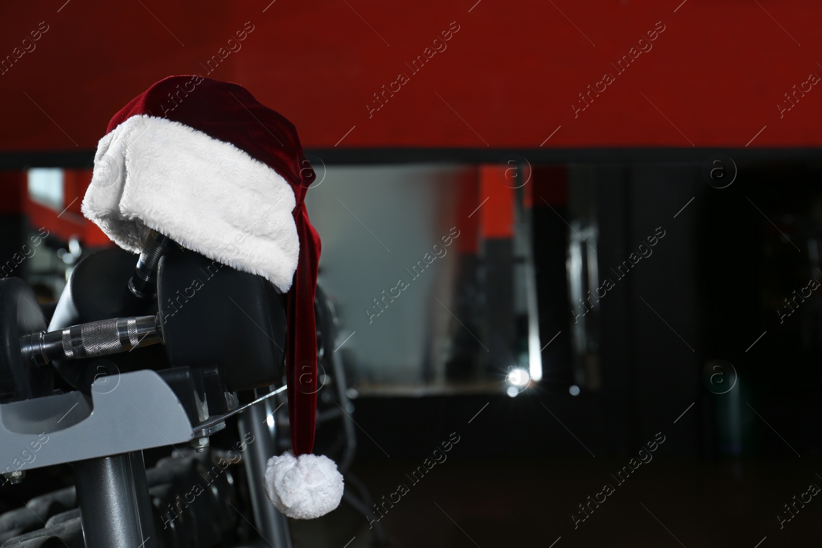 Photo of Santa hat on stand with dumbbells in modern gym. Space for text