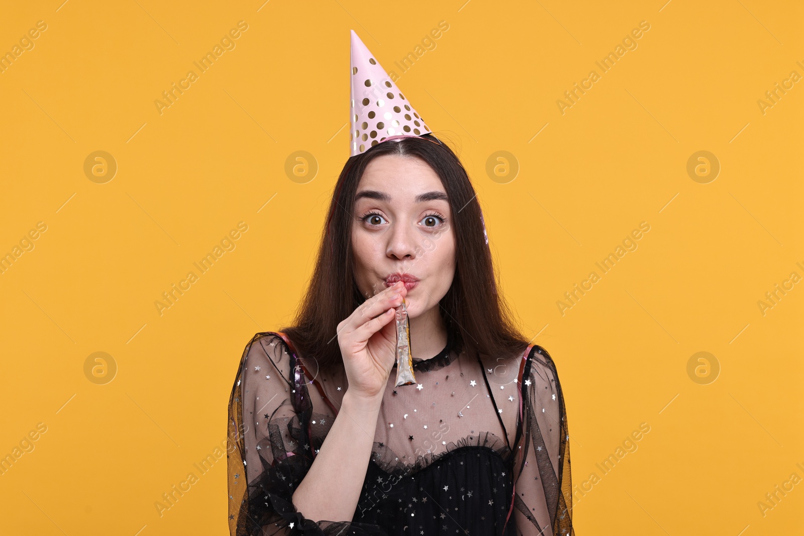 Photo of Woman in party hat with blower and streamers on orange background