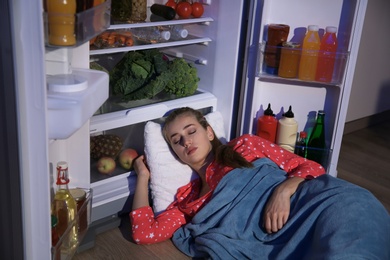 Photo of Young woman sleeping near refrigerator at night