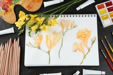Photo of Painting of freesias in sketchbook, flowers and art supplies on black table, flat lay