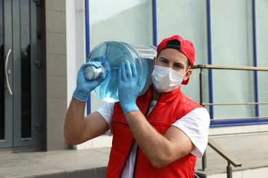 Photo of Courier in medical mask holding bottle for water cooler near building outdoors. Delivery during coronavirus quarantine