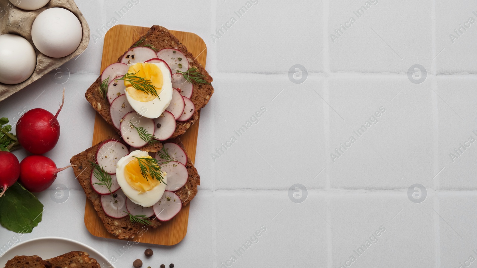 Photo of Tasty sandwiches with boiled egg, radish and ingredients on white tiled table, flat lay. Space for text