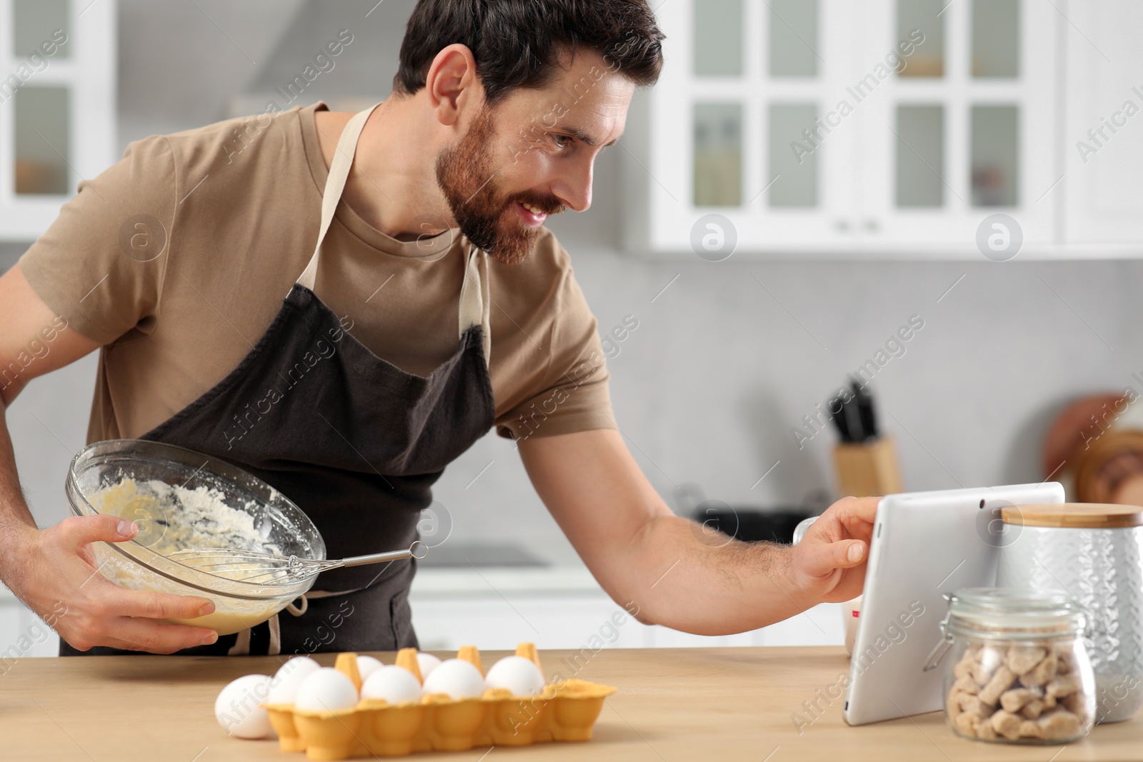 Photo of Man making dough while watching online cooking course via tablet in kitchen