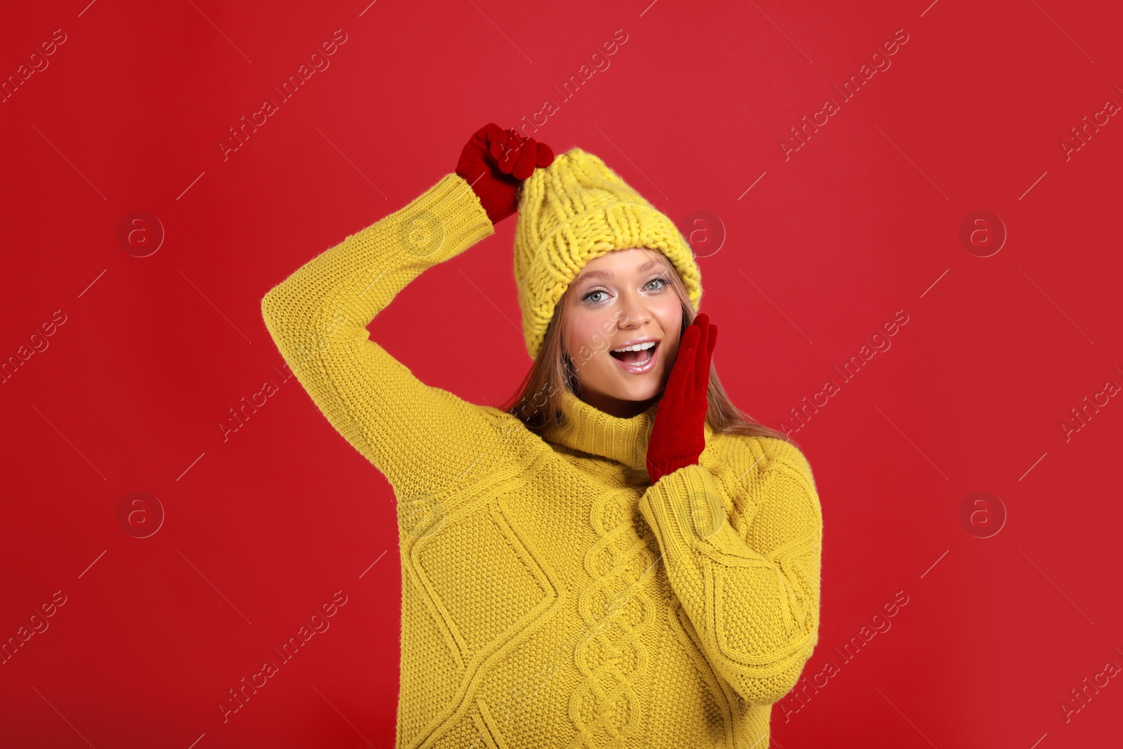 Photo of Emotional young woman in warm sweater, gloves and hat on red background. Winter season