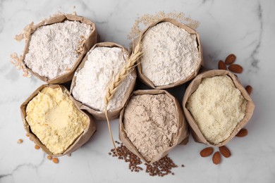 Photo of Paper bags with different types of flour and ingredients on white marble table, flat lay