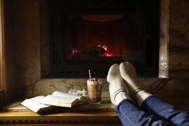 Photo of Woman with glass of hot cocoa and books near fireplace at home, closeup