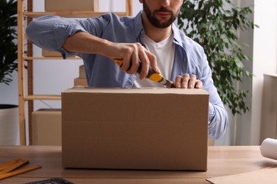 Photo of Man using utility knife to open parcel at wooden table indoors, closeup