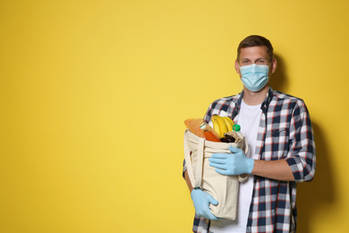 Photo of Male volunteer in protective mask and gloves with products on yellow background, space for text. Aid during coronavirus quarantine