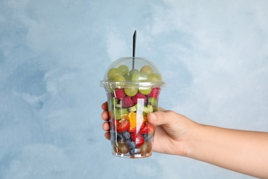 Photo of Woman holding plastic cup with fresh tasty fruit salad against light blue background, closeup