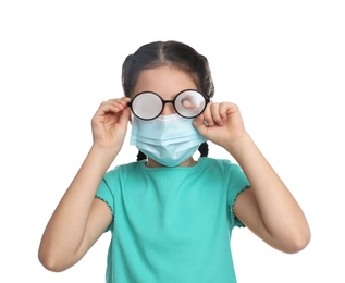 Photo of Little girl wiping foggy glasses caused by wearing medical face mask on white background. Protective measure during coronavirus pandemic