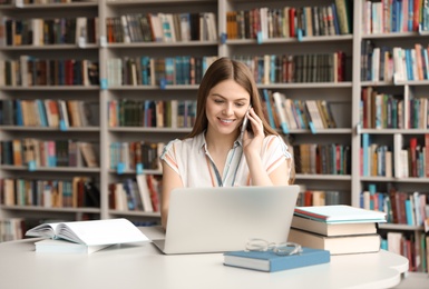 Photo of Young woman talking on phone and working with laptop at table in library