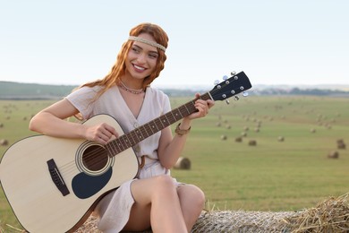 Photo of Beautiful hippie woman playing guitar on hay bale in field, space for text
