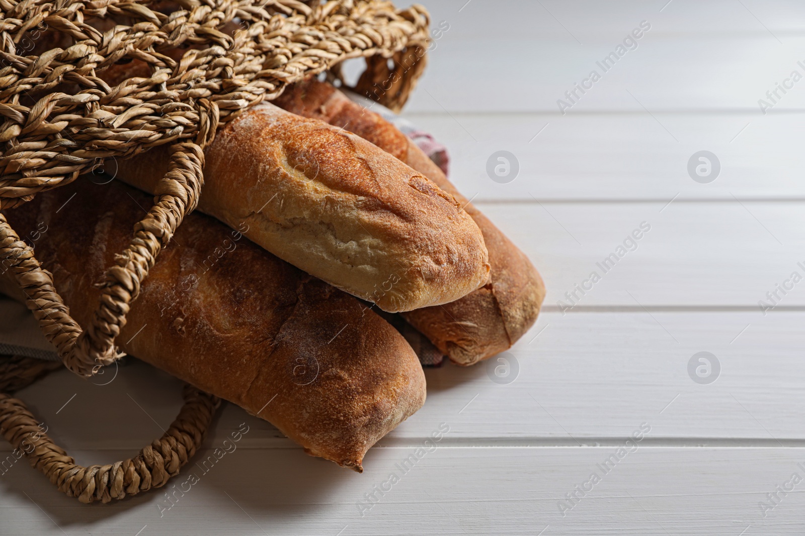 Photo of Wicker bag with fresh crispy ciabattas on white wooden table, closeup. Space for text