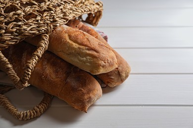 Photo of Wicker bag with fresh crispy ciabattas on white wooden table, closeup. Space for text