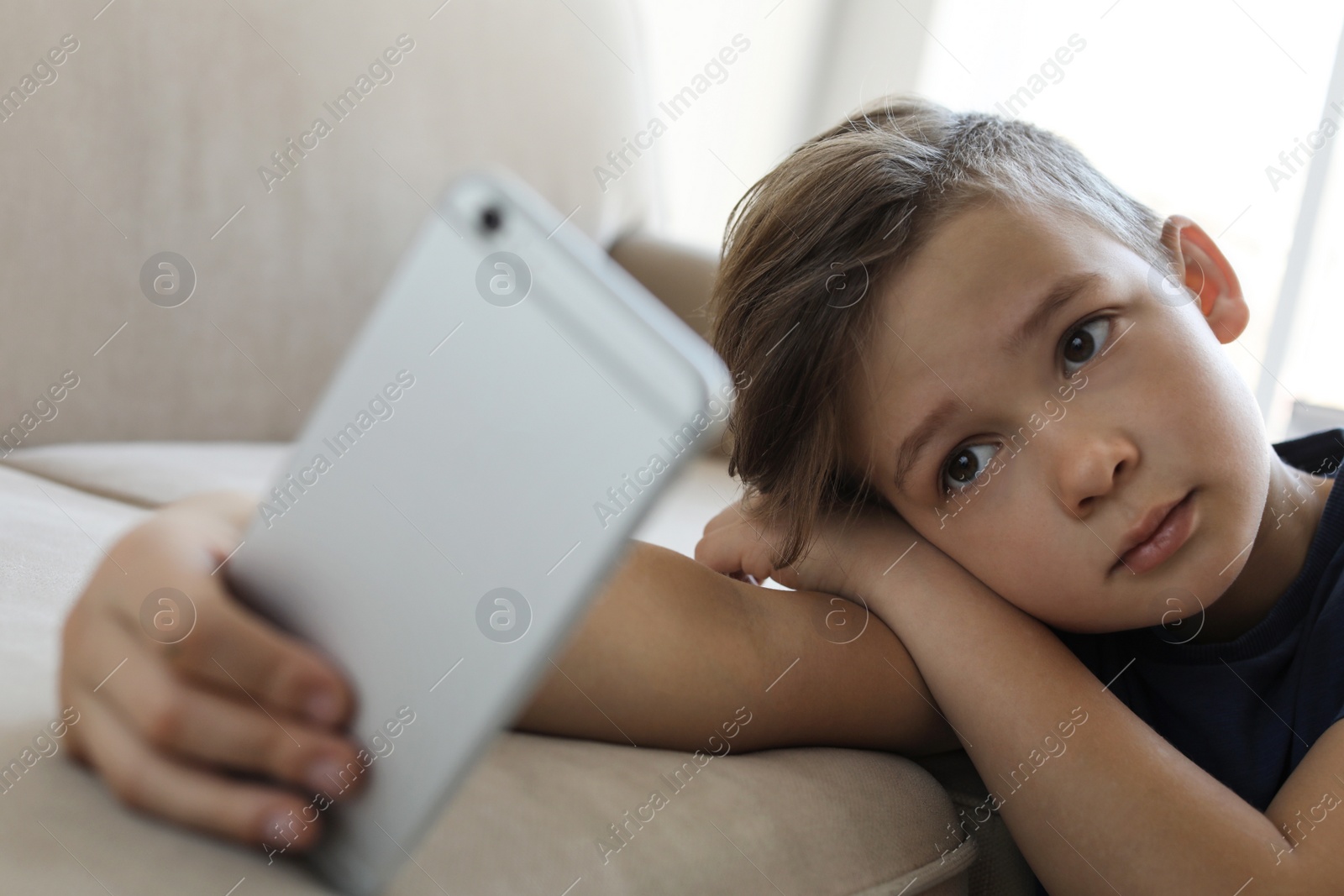 Photo of Sad little child with smartphone leaning on sofa in room. Danger of internet