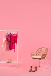Rack with different stylish women`s clothes, shoes and armchair on pink background