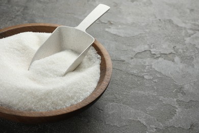 Photo of Granulated sugar in bowl and scoop on grey textured table, closeup. Space for text