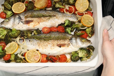 Photo of Woman holding baking dish with delicious fish and vegetables, top view