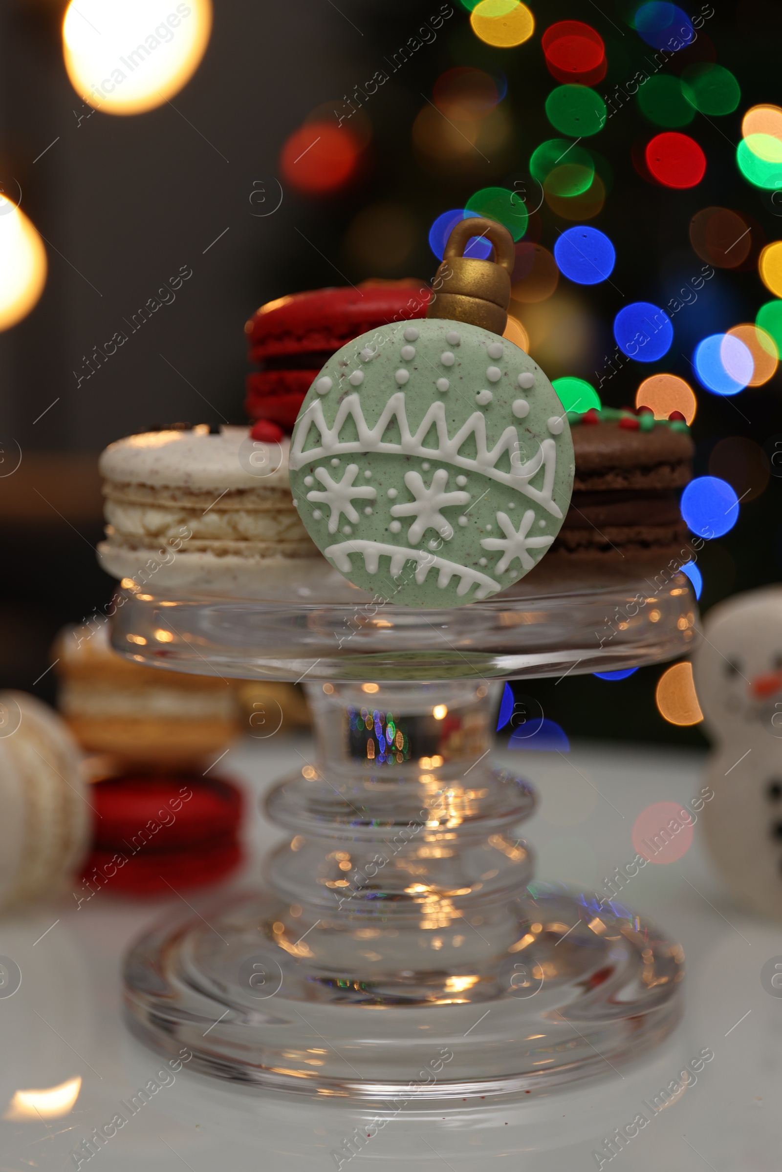 Photo of Beautifully decorated Christmas macarons on white table against blurred festive lights