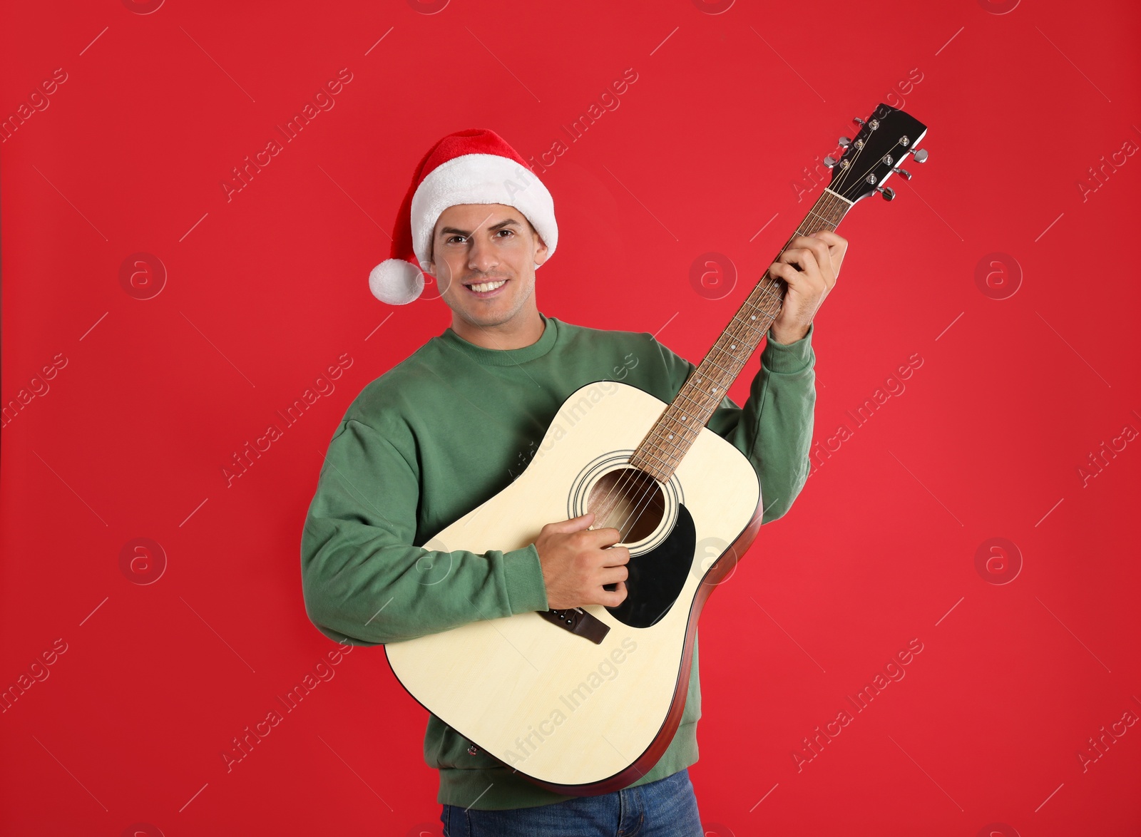 Photo of Man in Santa hat playing acoustic guitar on red background. Christmas music