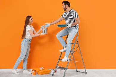Woman giving can of paint to man near orange wall indoors. Interior design