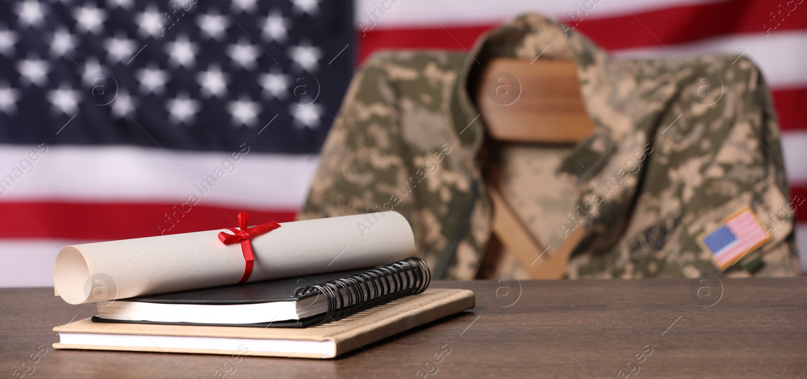 Image of Military education. Notebooks and diploma on wooden table, chair with soldier's jacket against flag of United States indoors