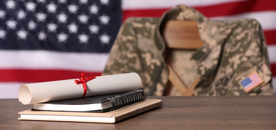 Image of Military education. Notebooks and diploma on wooden table, chair with soldier's jacket against flag of United States indoors