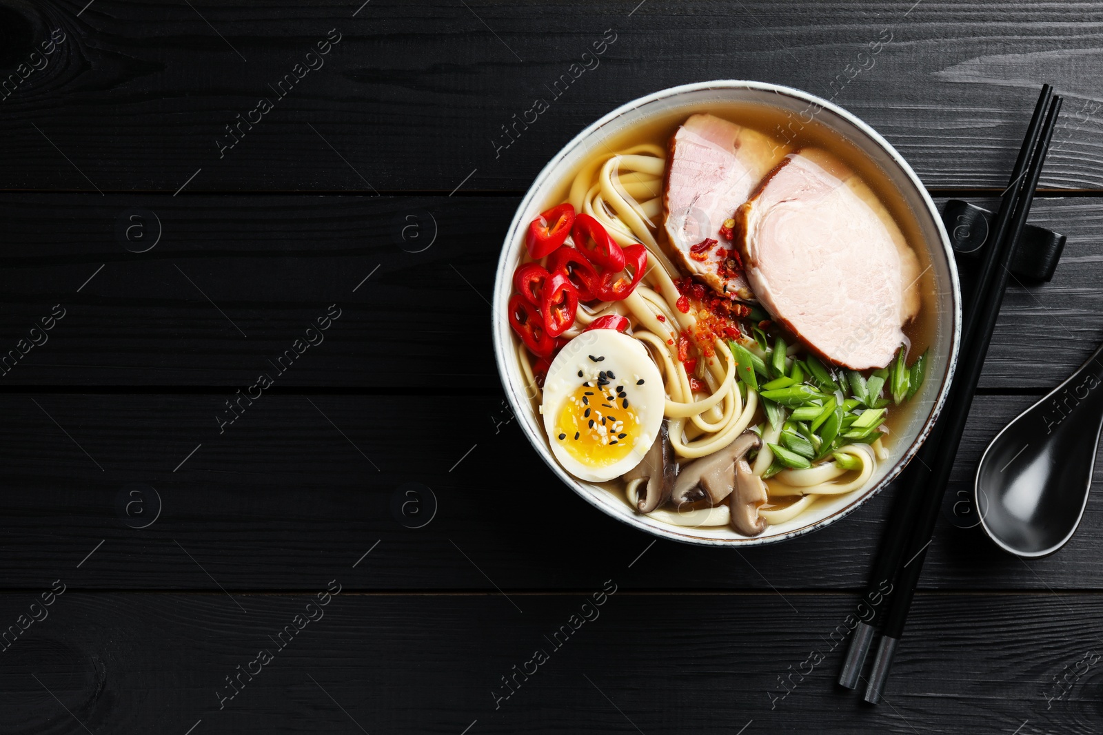 Photo of Delicious ramen in bowl, spoon and chopsticks on black wooden table, flat lay with space for text. Noodle soup