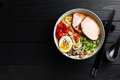 Photo of Delicious ramen in bowl, spoon and chopsticks on black wooden table, flat lay with space for text. Noodle soup