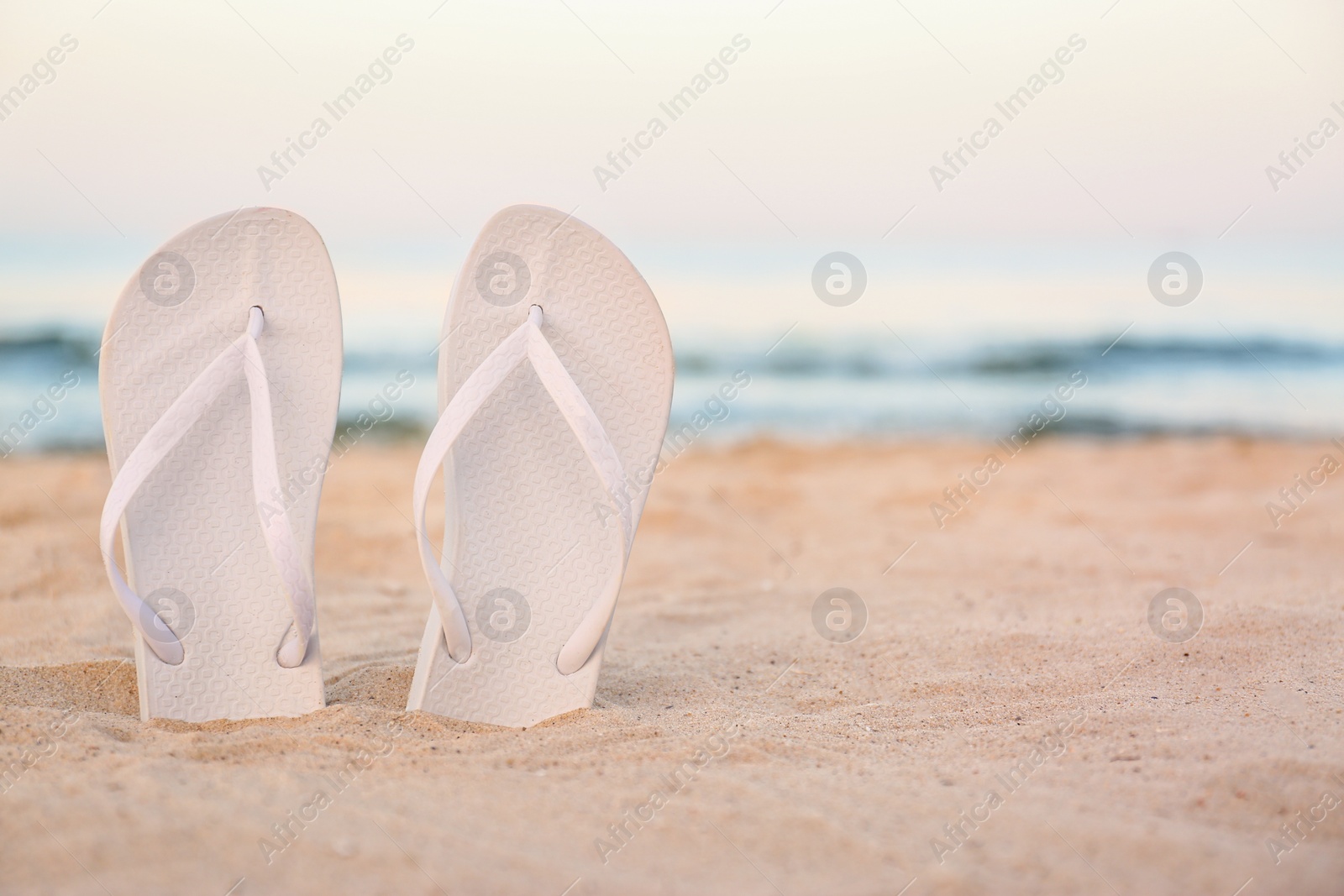Photo of White flip flops on sand near sea, space for text. Beach accessories