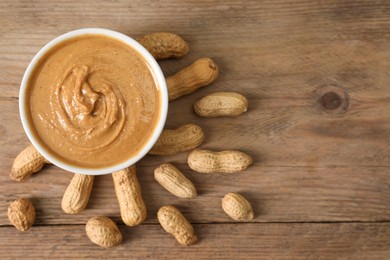 Photo of Delicious nut butter in bowl and peanuts on wooden table, top view. Space for text