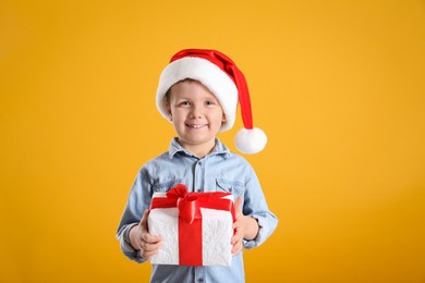 Photo of Cute little boy in Santa Claus hat holding gift box on yellow background