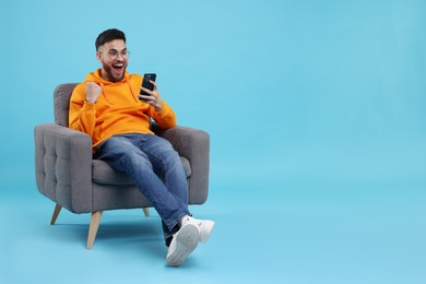 Photo of Happy young man using smartphone on armchair against light blue background, space for text