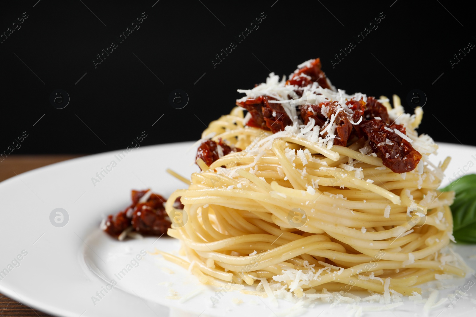 Photo of Tasty spaghetti with sun-dried tomatoes and parmesan cheese on plate against black background, closeup. Exquisite presentation of pasta dish