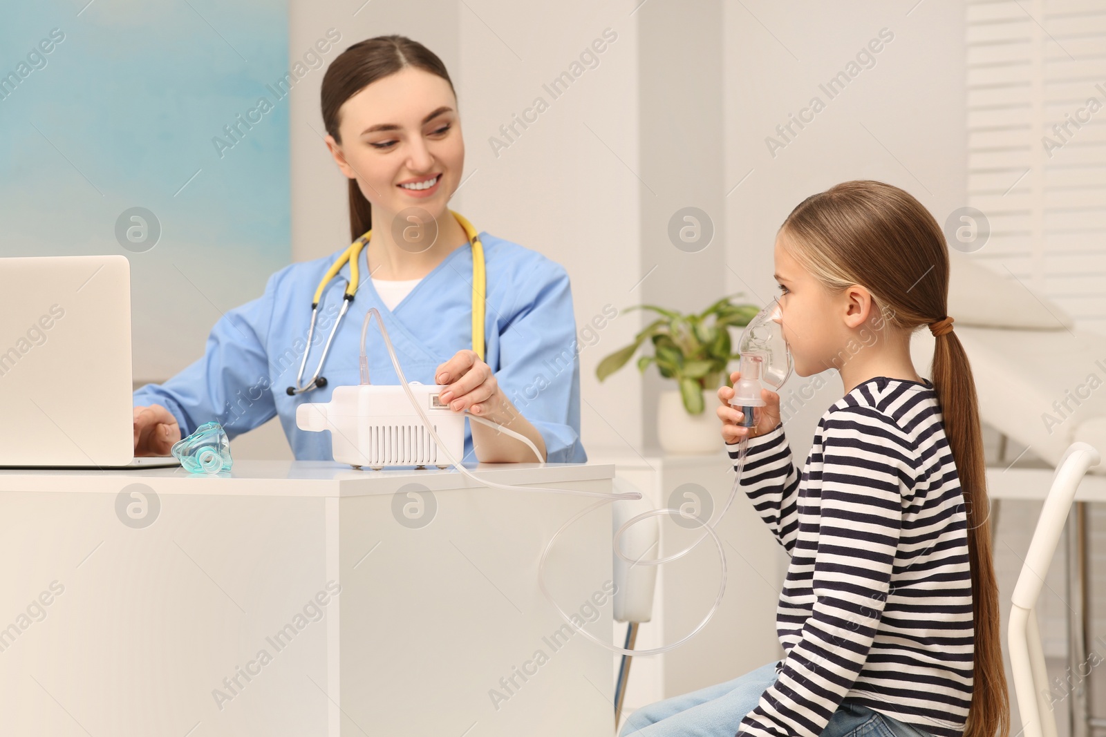 Photo of Medical assistant sitting near sick little girl while she using nebulizer for inhalation in hospital