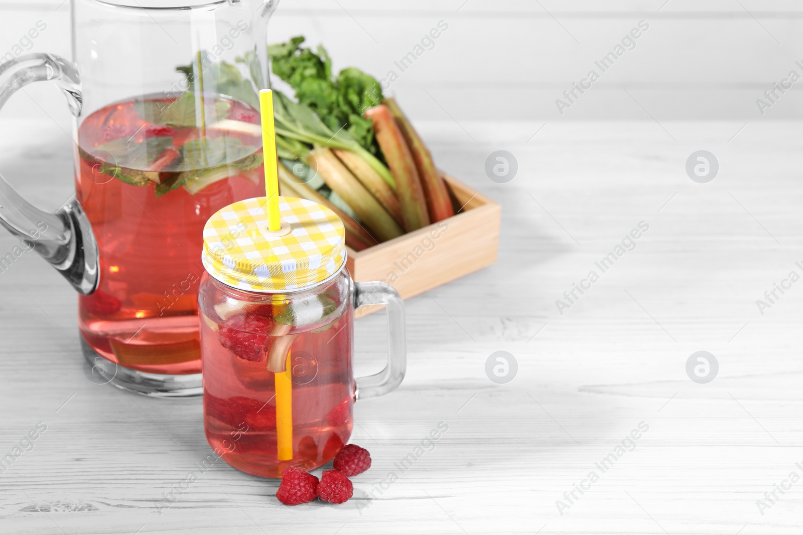 Photo of Mason jar and jug of tasty rhubarb cocktail with raspberry on white wooden table, space for text