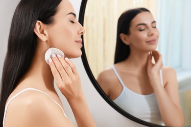 Photo of Young woman using cotton pad with micellar water near mirror indoors