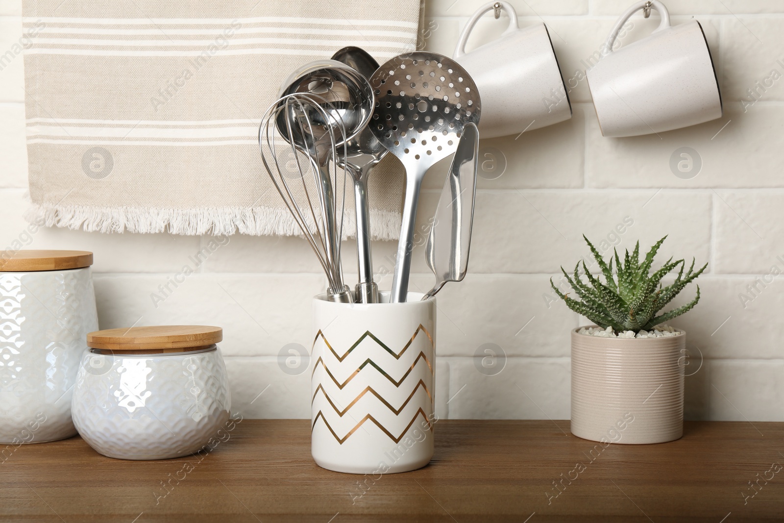 Photo of Steel utensils and different dishware on wooden table near white brick wall in kitchen