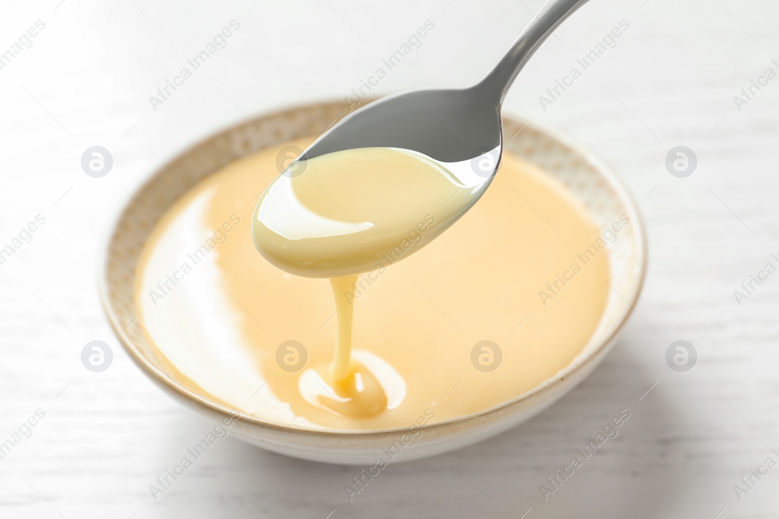 Photo of Spoon with pouring condensed milk over bowl on white background, closeup. Dairy products