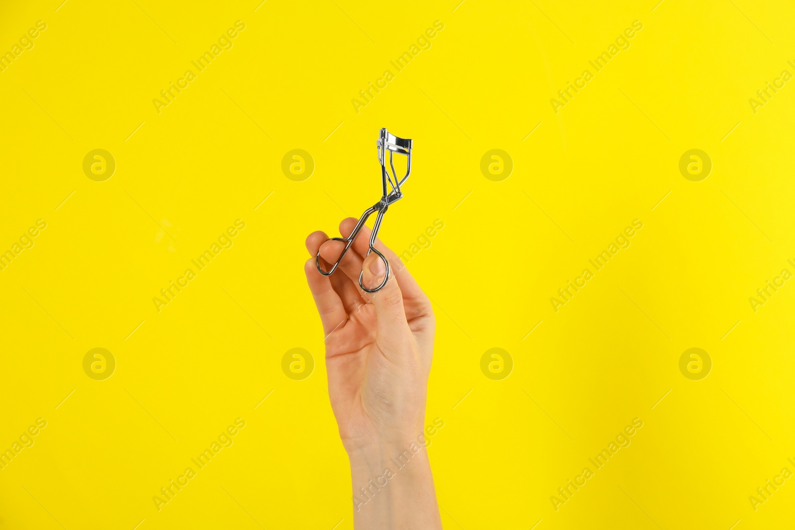 Photo of Woman holding eyelash curler on yellow background, closeup. Makeup tool