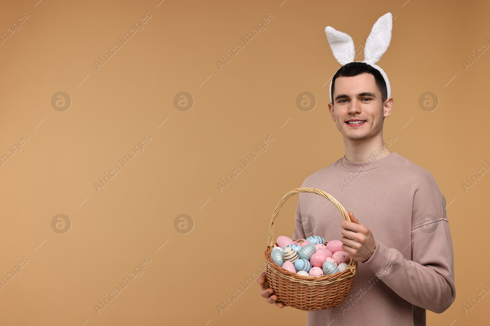 Photo of Easter celebration. Handsome young man with bunny ears holding basket of painted eggs on beige background. Space for text