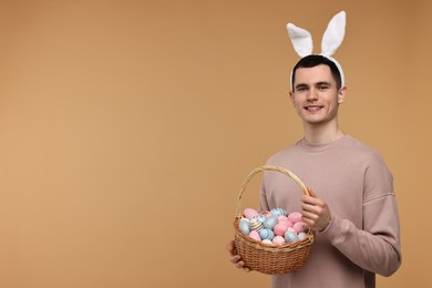 Easter celebration. Handsome young man with bunny ears holding basket of painted eggs on beige background. Space for text